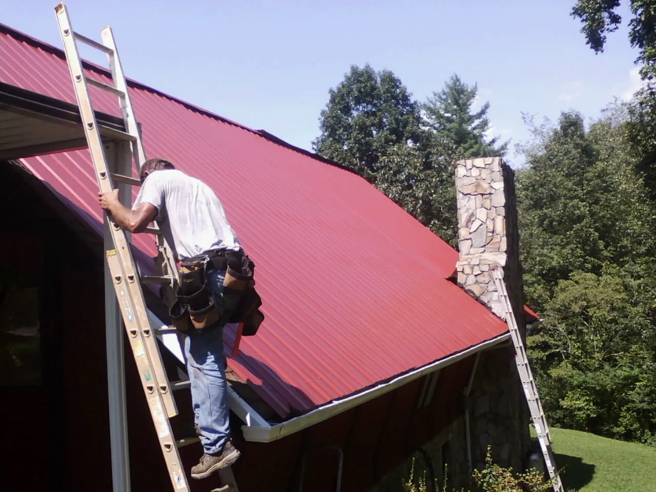 Roofer on a ladder beside a red metal roof and stone chimney — Soffit & Fascia Repair by Roofing Brothers in Indianapolis